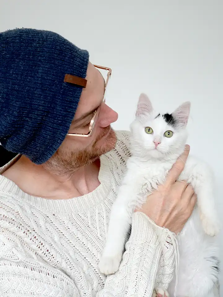 Nous pouvons voir Valentin Duchenne, fondateur de Pilea Studio et le webdesigner du site des amis d'Ivar tenir un chat blanc et noir. Valentin qui regarde le chat est assis sur une chaise de bureau avec un pull blanc et un bonnet bleu. Il tient dans ses mains Miaouss un chat à poils longs.